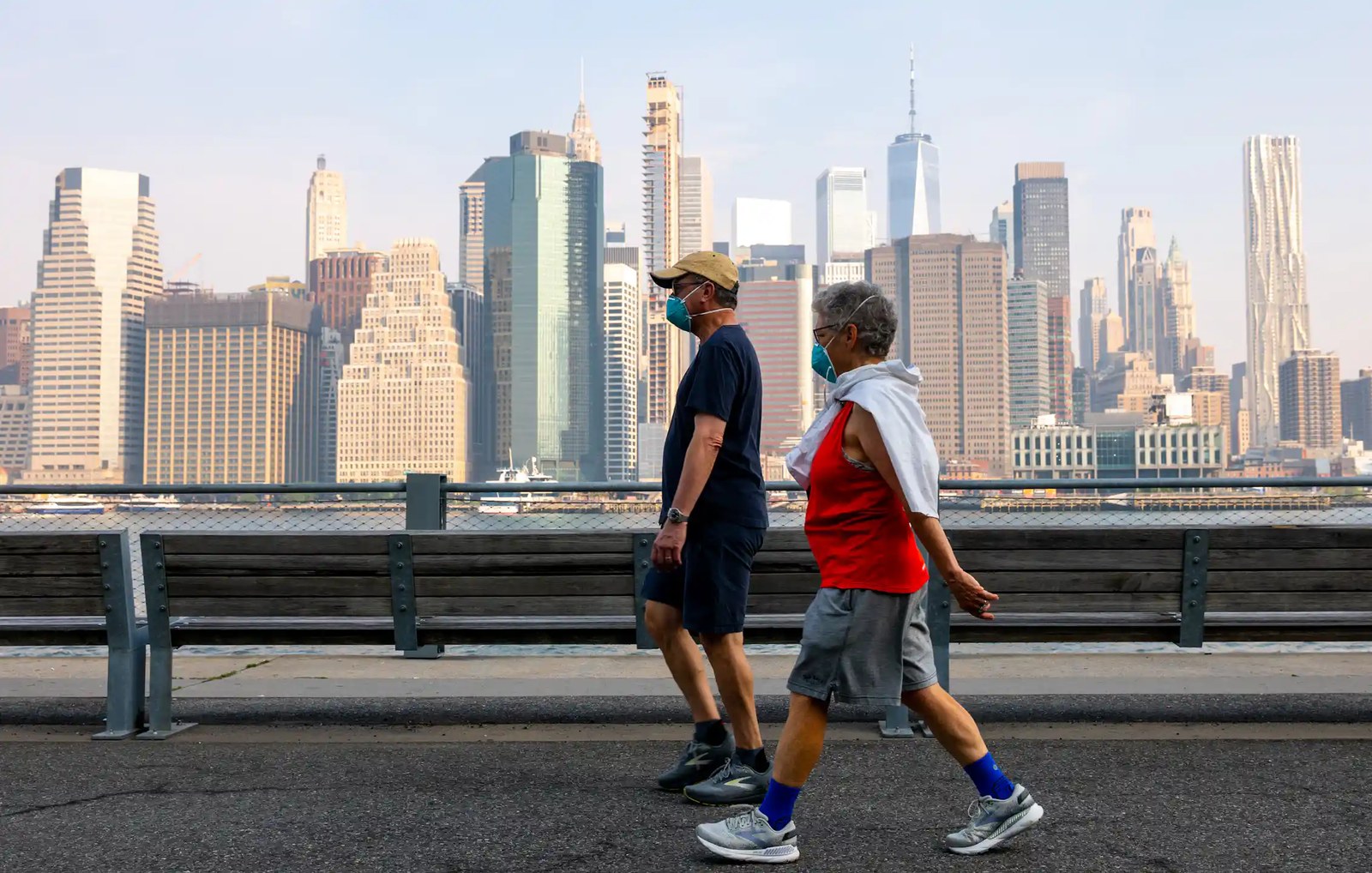 Two people wear masks over their nose and mouth while they walk along a path with a cityscape visible in the background.