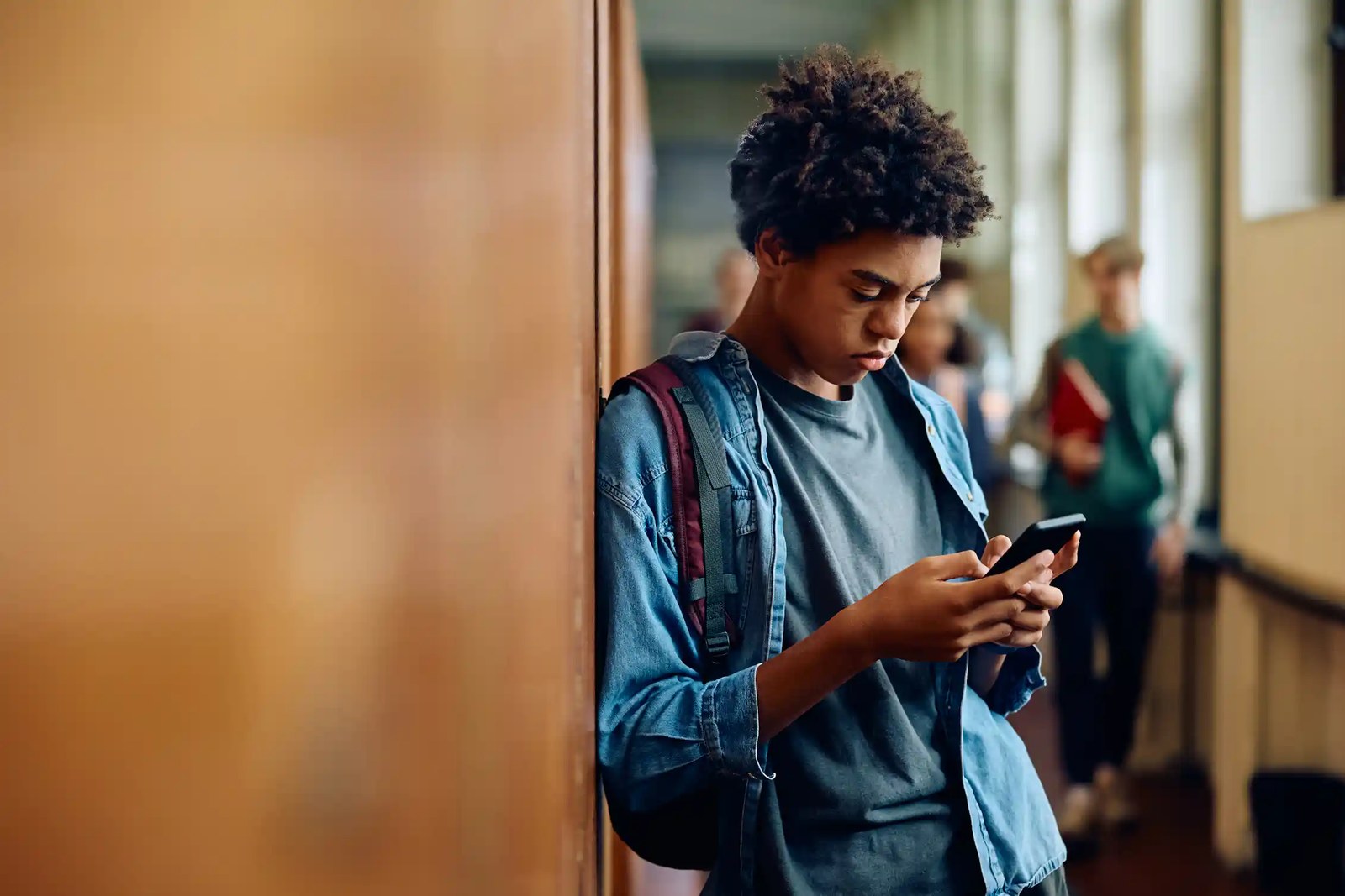 a teen boy leans against a wall while looking at a phone